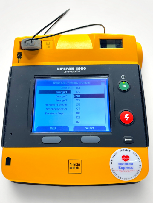 A Physio Control Lifepak 1000 AED defibrillator with a graphical/ECG display, orange casing, and a carrying case.