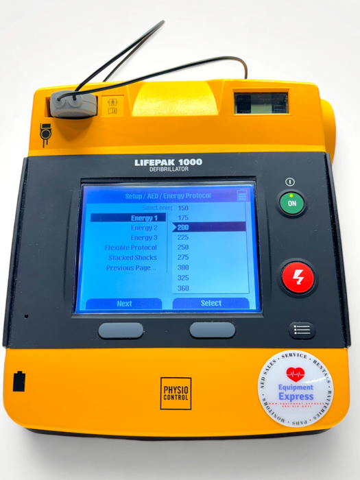 A Physio Control Lifepak 1000 AED defibrillator with a graphical/ECG display, orange casing, and a carrying case.